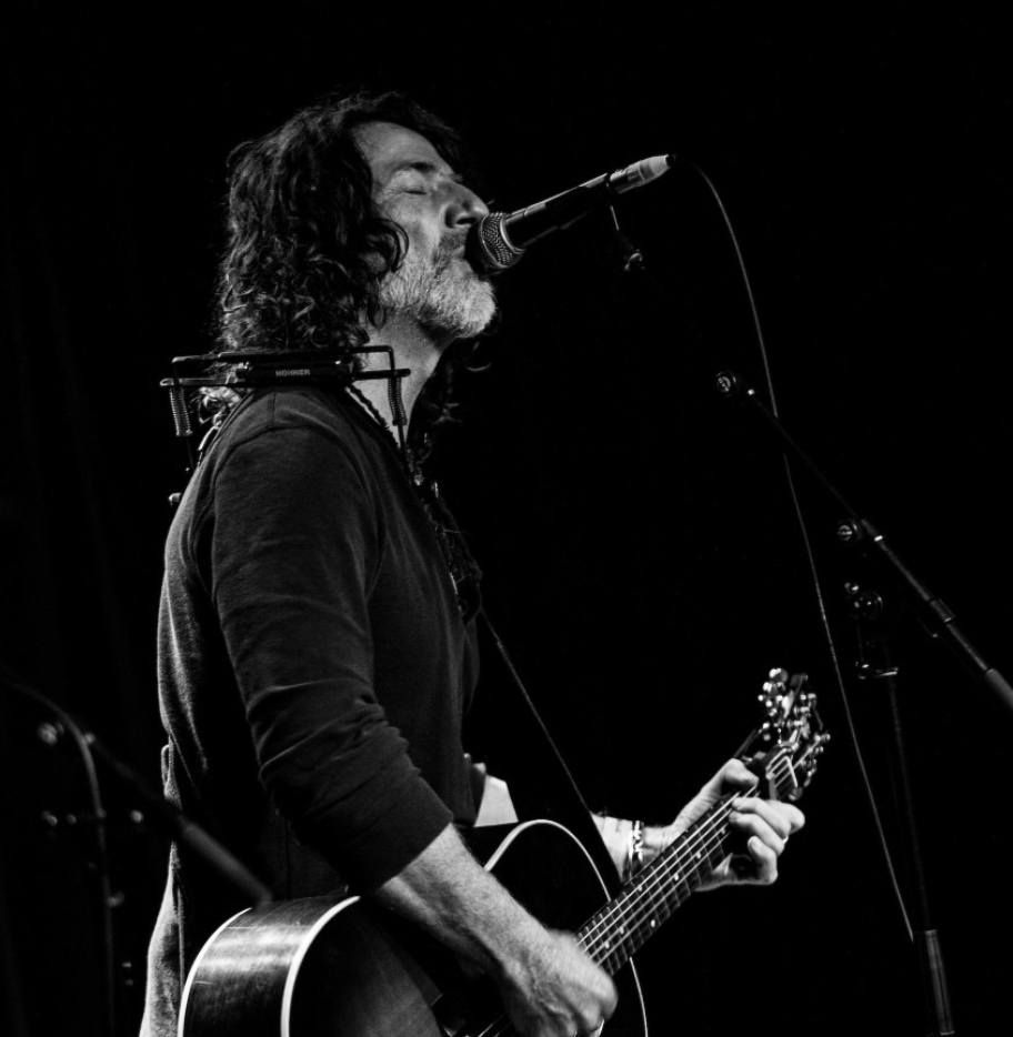Musician with long hair plays guitar and sings into a microphone on stage in a black and white image.